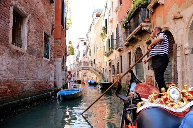 A gondolier in a striped shirt rowing a gondola down a narrow canal between brick houses with flower boxes