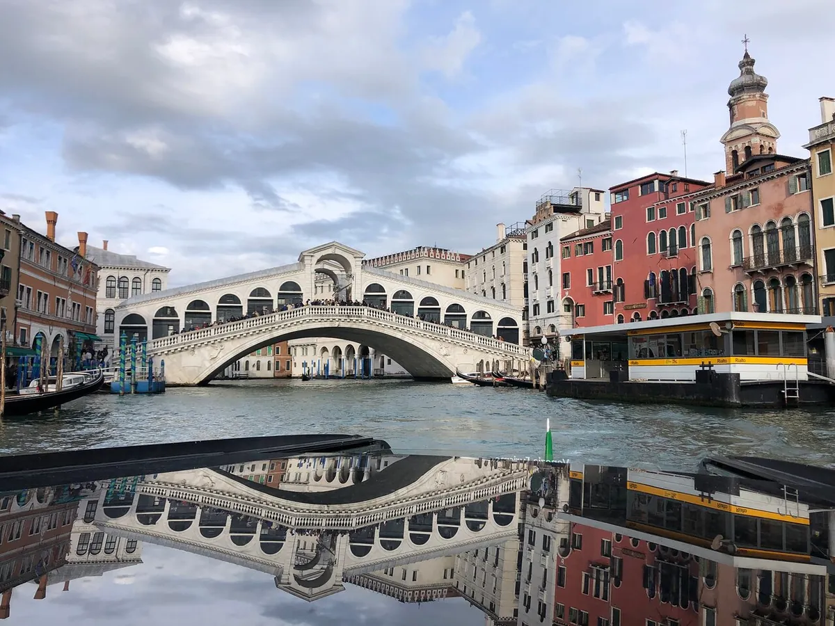 The white stone arch of the Rialto Bridge reflected in the still Grand Canal, vaporetto pontoons on the right